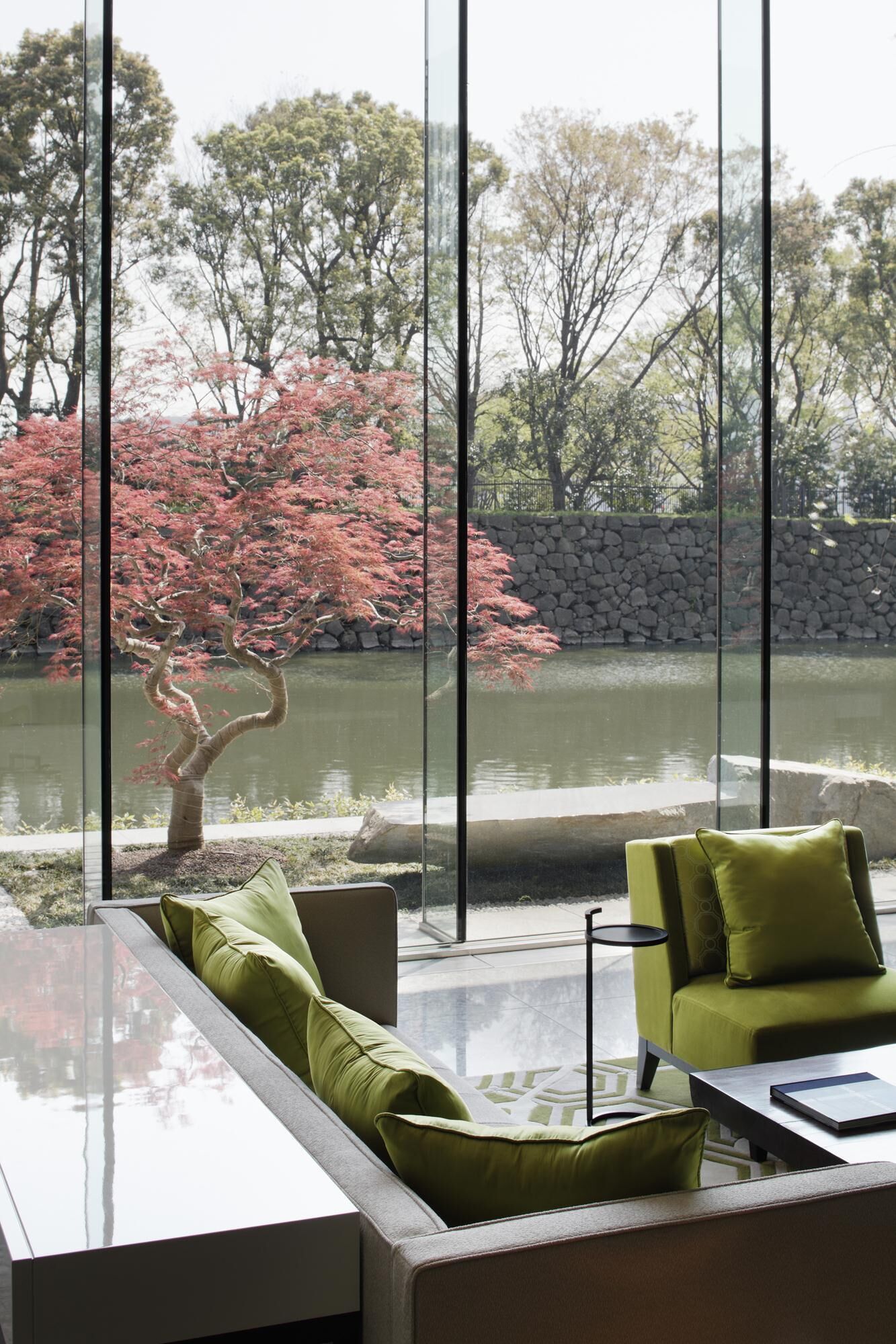 A modern interior with a glass table, green sofa, and a view of a pond with red foliage.