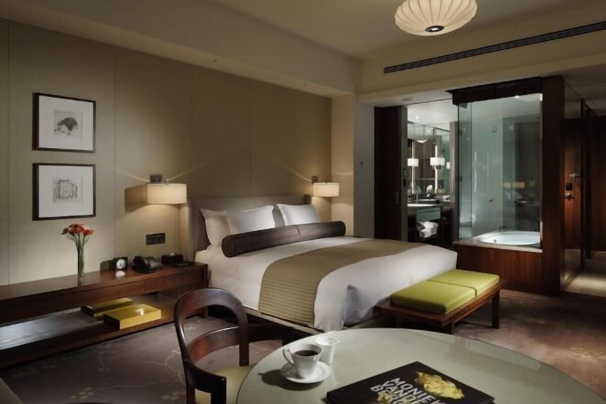 A hotel room with a bed, bedside tables, a chair, a round table with a cup and a menu, and a bathroom visible through an open door.