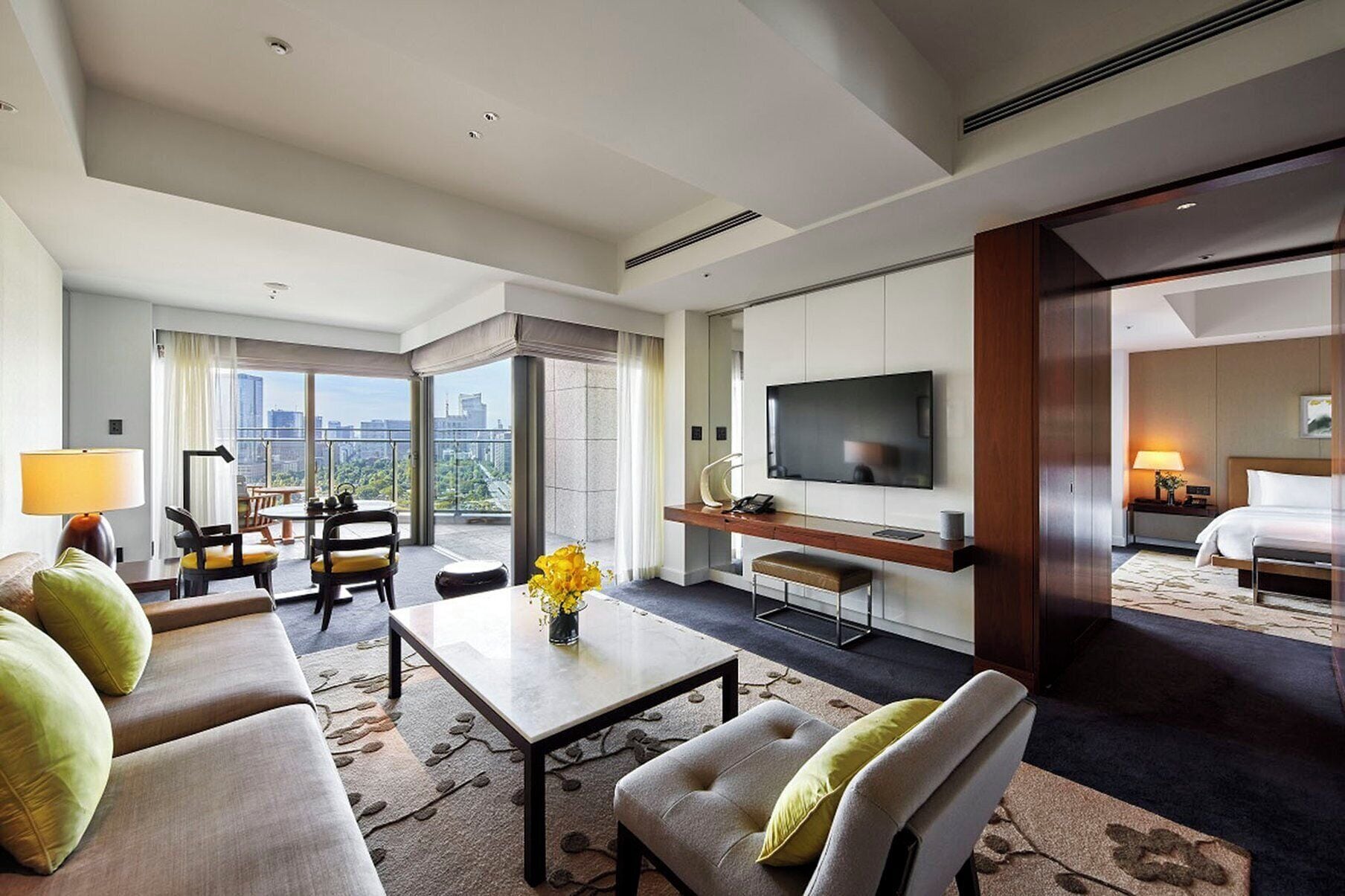 A modern hotel room with a sofa, a coffee table, a TV, and a balcony with a city view.