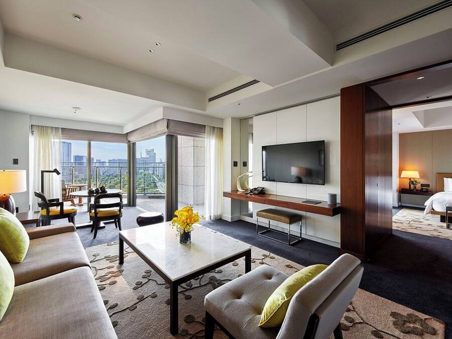 A modern hotel room with a sofa, a coffee table, a TV, and a balcony with a city view.