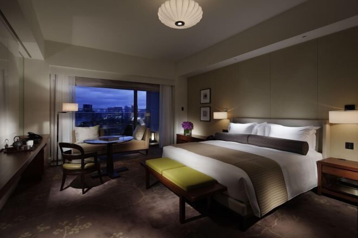 A hotel room with a large bed, a desk, a chair, and a view of the city at dusk.