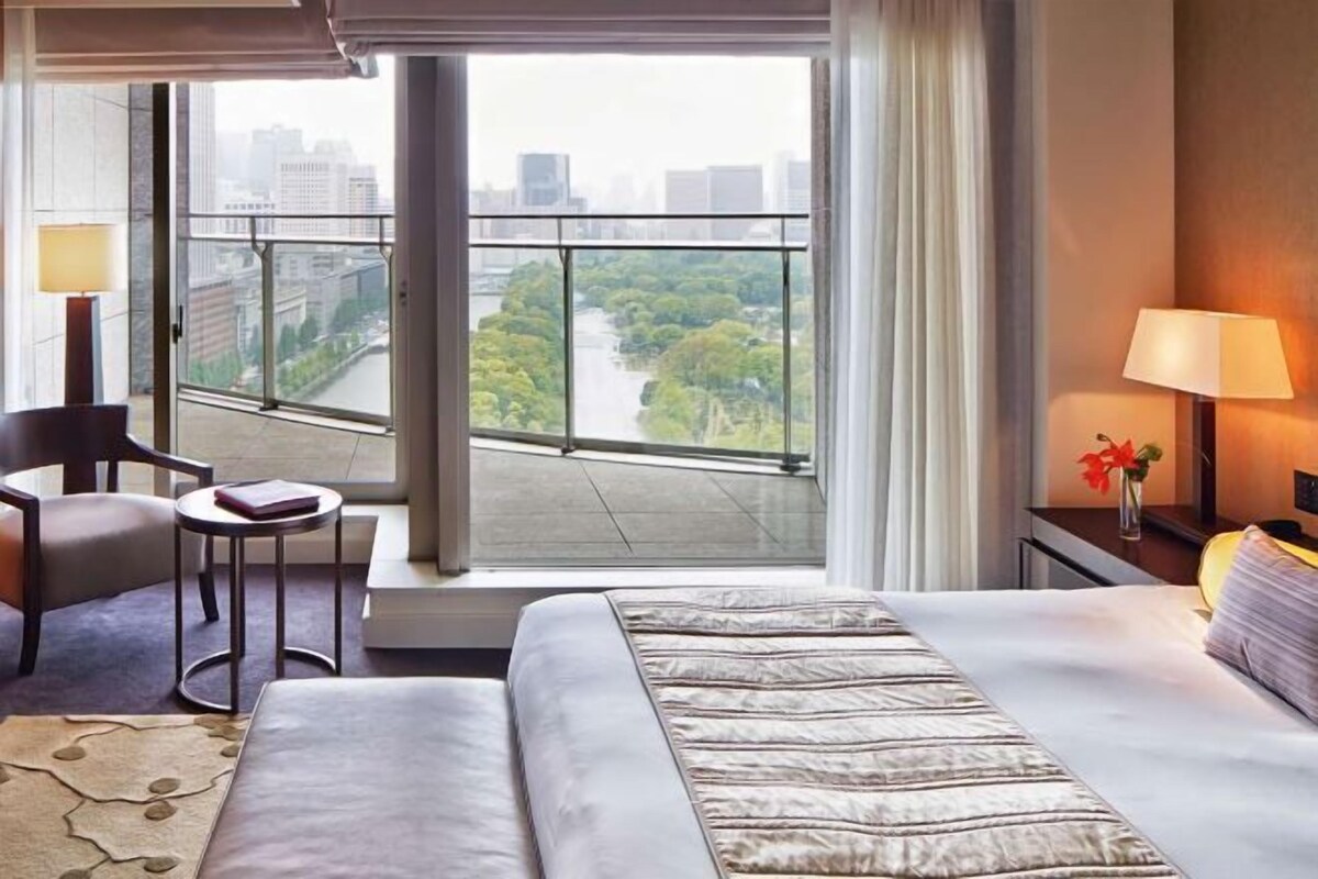 A hotel room with a large window offering a view of a river and cityscape. There is a bed with a patterned bedspread, a chair, a small round table, and a nightstand with a lamp.