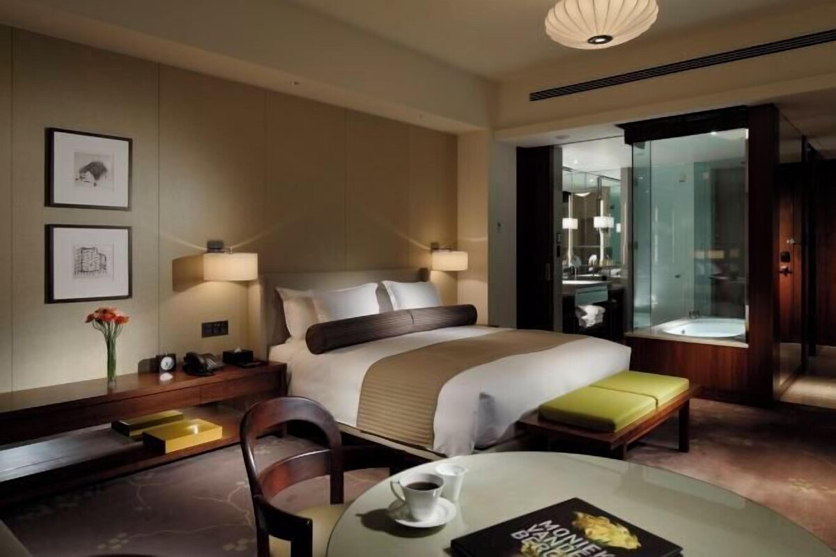 A hotel room with a large bed, bedside lamps, a desk with a chair, a glass table with a cup and magazine, and a bathroom visible through a glass door.