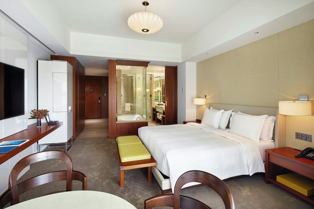 A hotel room with a bed, bedside table, lamp, desk, chair, and a bathroom visible through an open door.