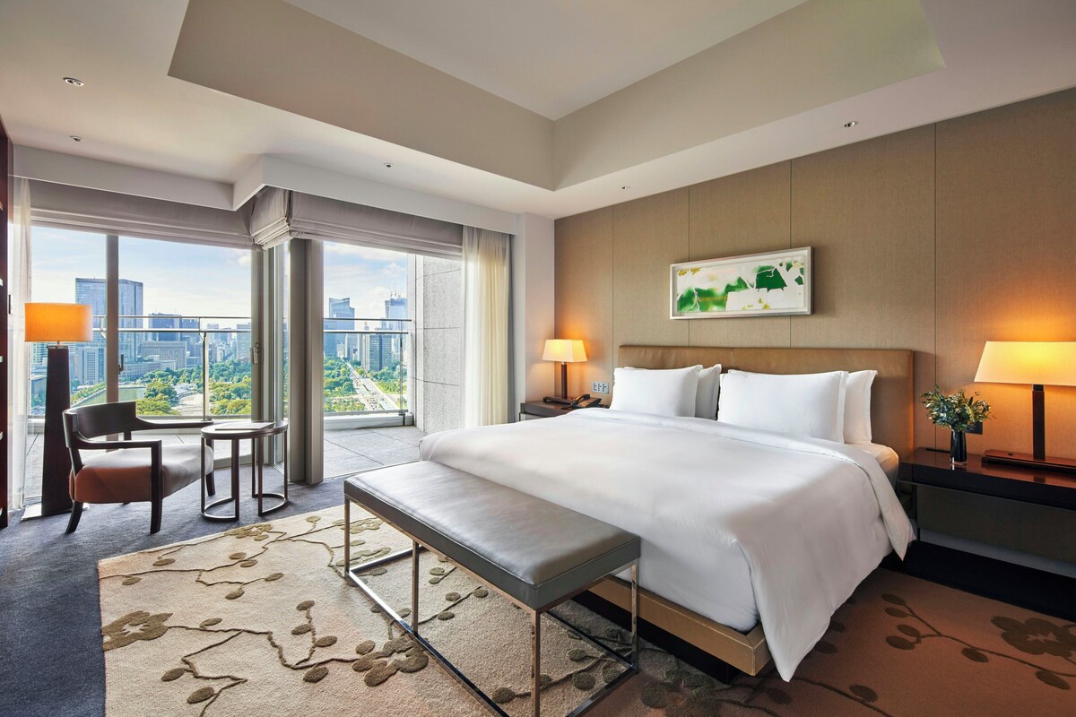A hotel room with a large bed, a desk, a chair, and a view of the city.