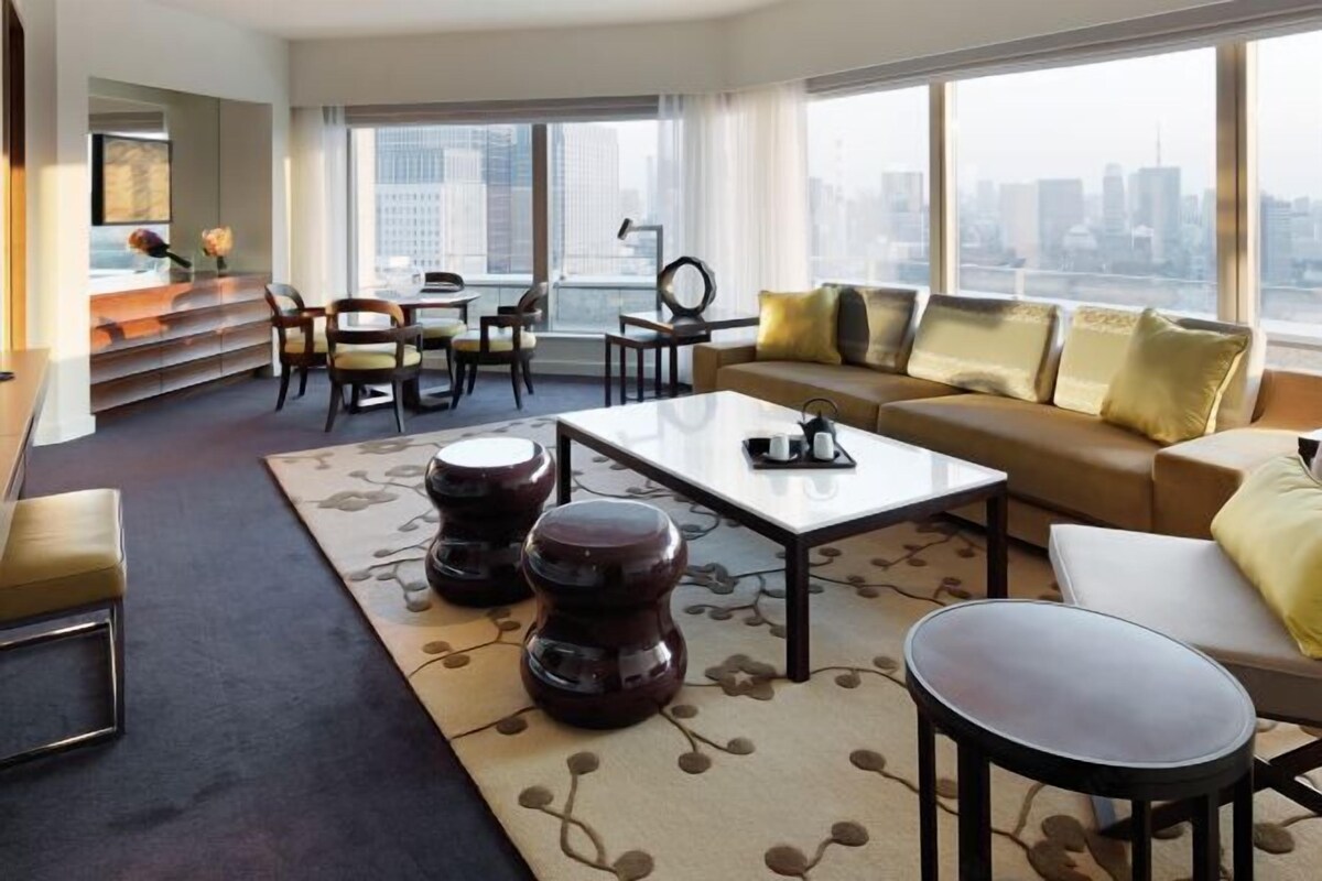 A modern hotel room with a large window offering a city view, a sofa set, a dining area, and a small table with a decorative item.