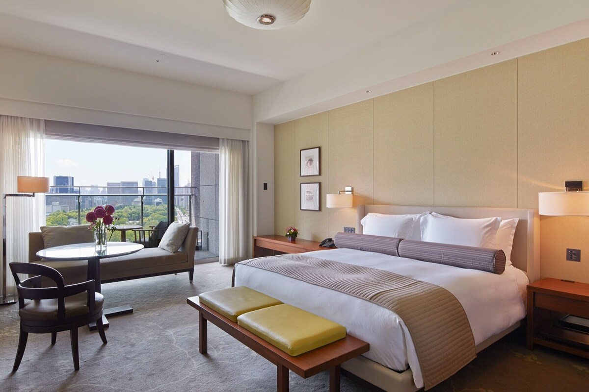 A hotel room with a bed, bedside tables, a desk, a chair, a small table with flowers, and a view of the city through large windows.