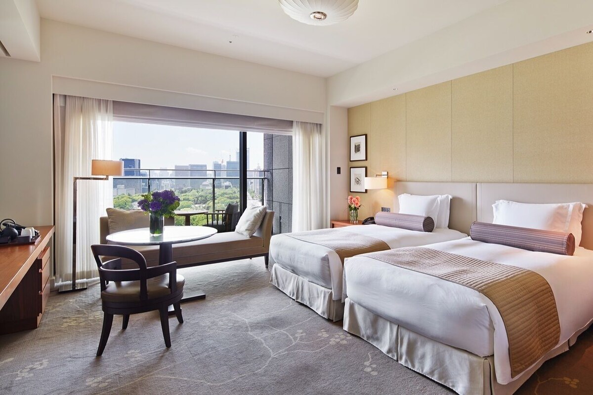 A hotel room with two beds, a desk, a chair, a round table with a vase, and a view of the city.