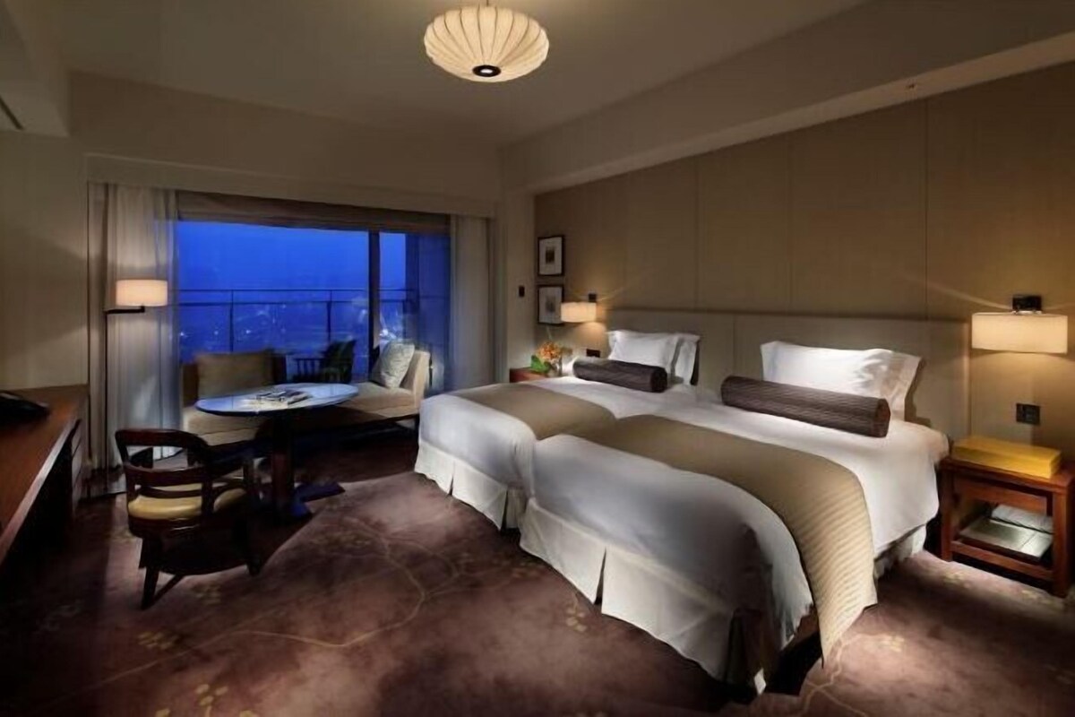 A hotel room with two beds, a desk, a chair, and a view of the city at dusk.