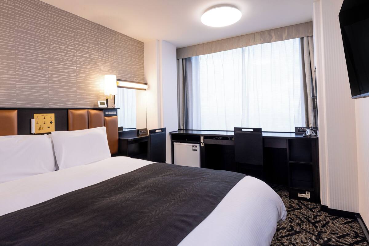 Ett hotellrum med en stor säng, ett skrivbord, en stol, en platt-TV och ett fönster med gardiner.
