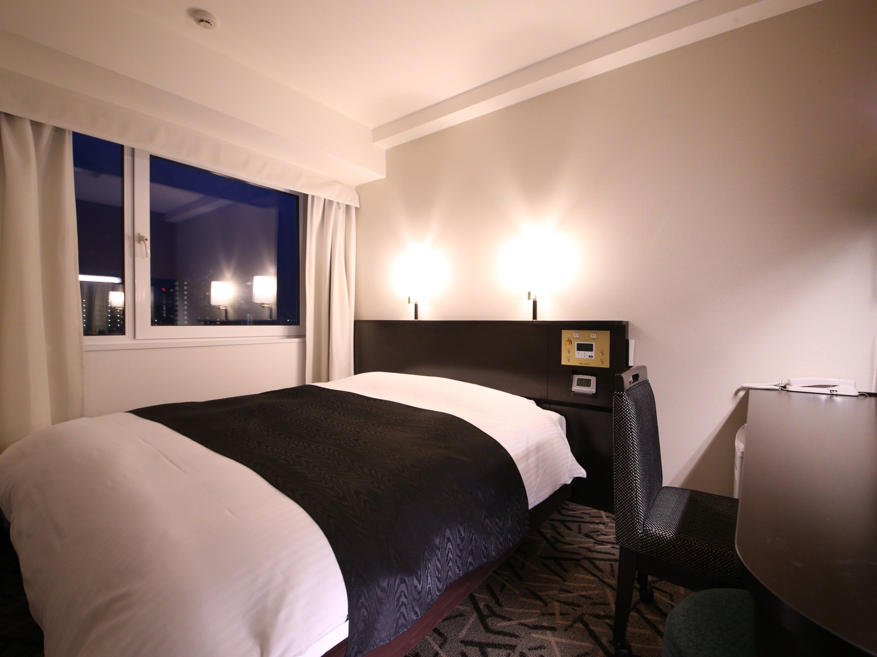 Een hotelkamer met een groot bed, een bureau met stoel, een raam met gordijnen en een bedieningspaneel aan de muur.