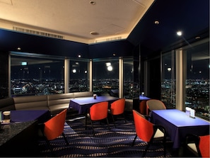 A modern hotel room with a city view, large windows, and a seating area with tables and chairs.