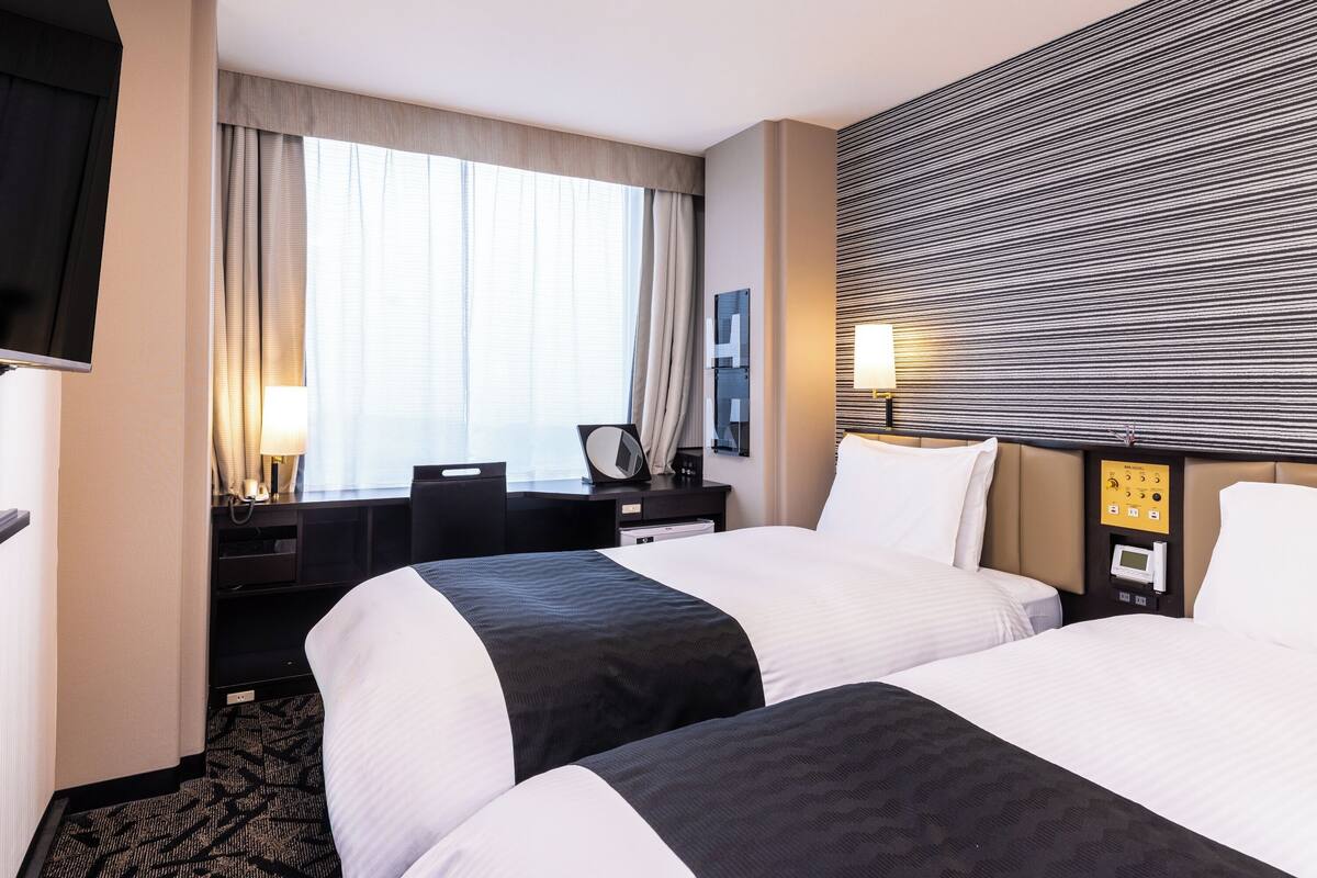 Ett hotellrum med två sängar, ett skrivbord med en stol, en TV och en väggmonterad kontrollpanel.