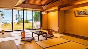 A traditional Japanese room with tatami flooring, a low table, two chairs, and a large window offering a view of a garden and water. - Hotel Ranpu (Hirado-shi)