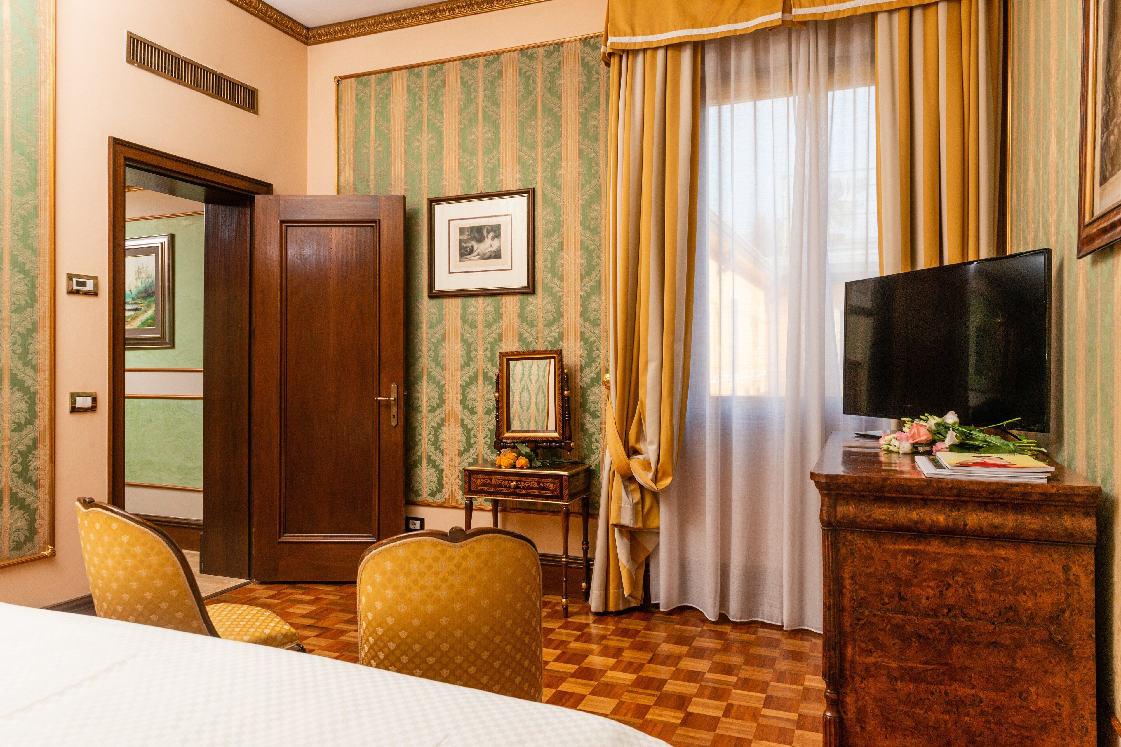 A hotel room with a wooden floor, a bed, a TV on a wooden stand, a desk with a mirror, and a chair.