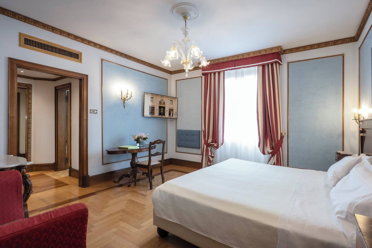 A hotel room with a large bed, a desk with a chair, a mirror, and a chandelier.