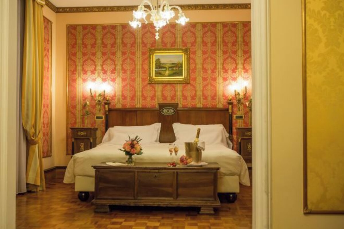 Una habitación de hotel con una cama grande, cabecera de madera, dos mesitas de noche con lámparas, una mesita auxiliar y un aparador de madera, además de una araña.