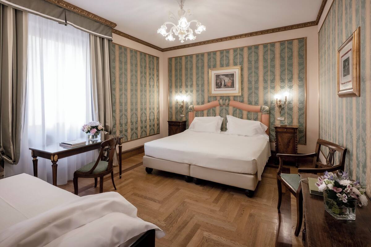 A hotel room with a large bed, a desk with a chair, a chandelier, and a painting on the wall.