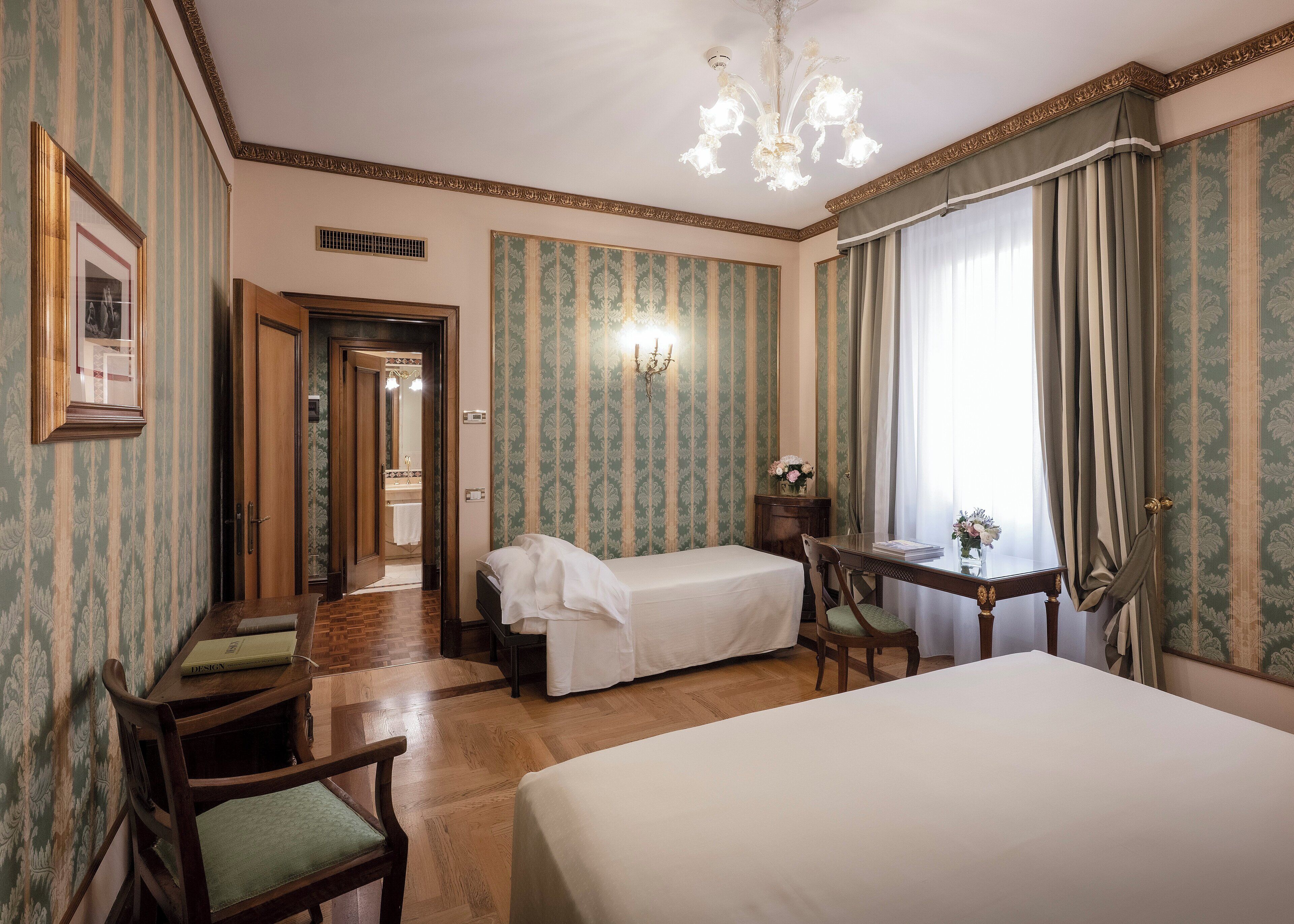 A hotel room with a large bed, a smaller bed, a desk with a chair, a chandelier, and patterned wallpaper.