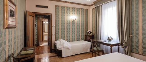 A hotel room with a large bed, a smaller bed, a desk with a chair, a chandelier, and patterned wallpaper.