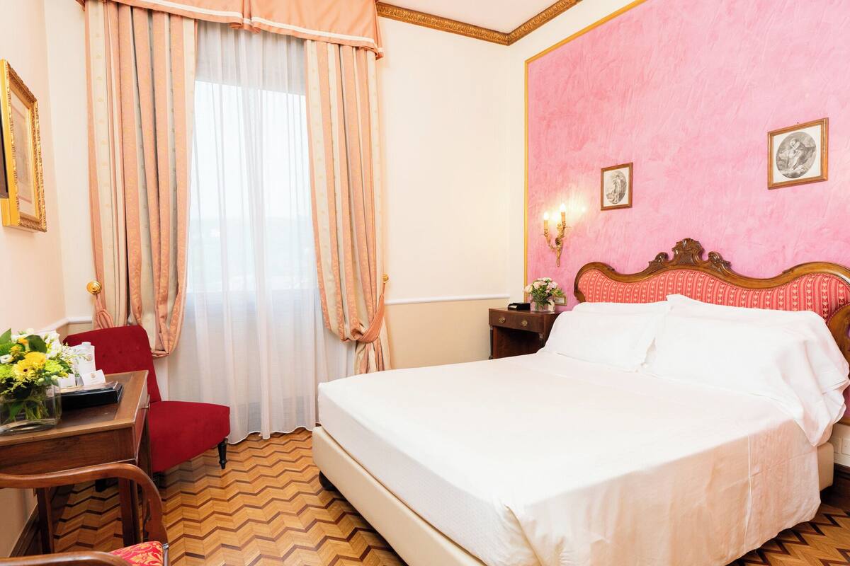A hotel room with a large bed, a desk with a chair, a red armchair, and a window with curtains.