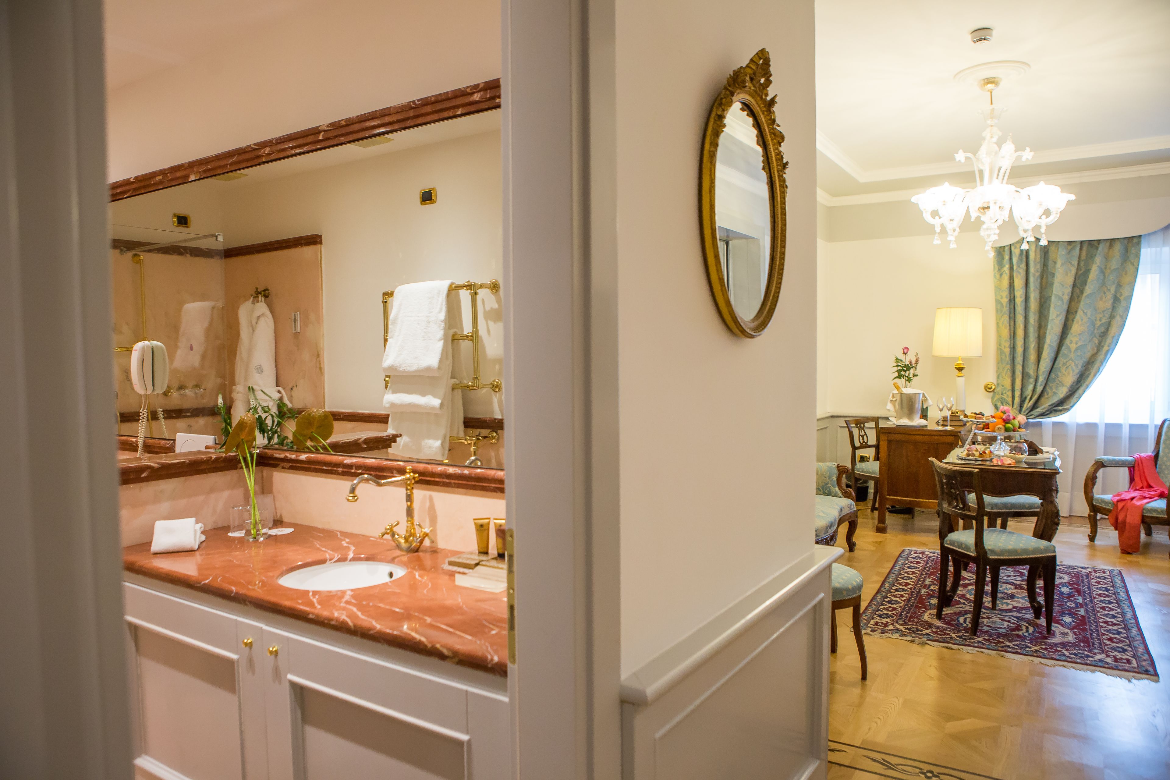 A bathroom with a marble sink, a large mirror, and a chandelier in the adjacent living area.