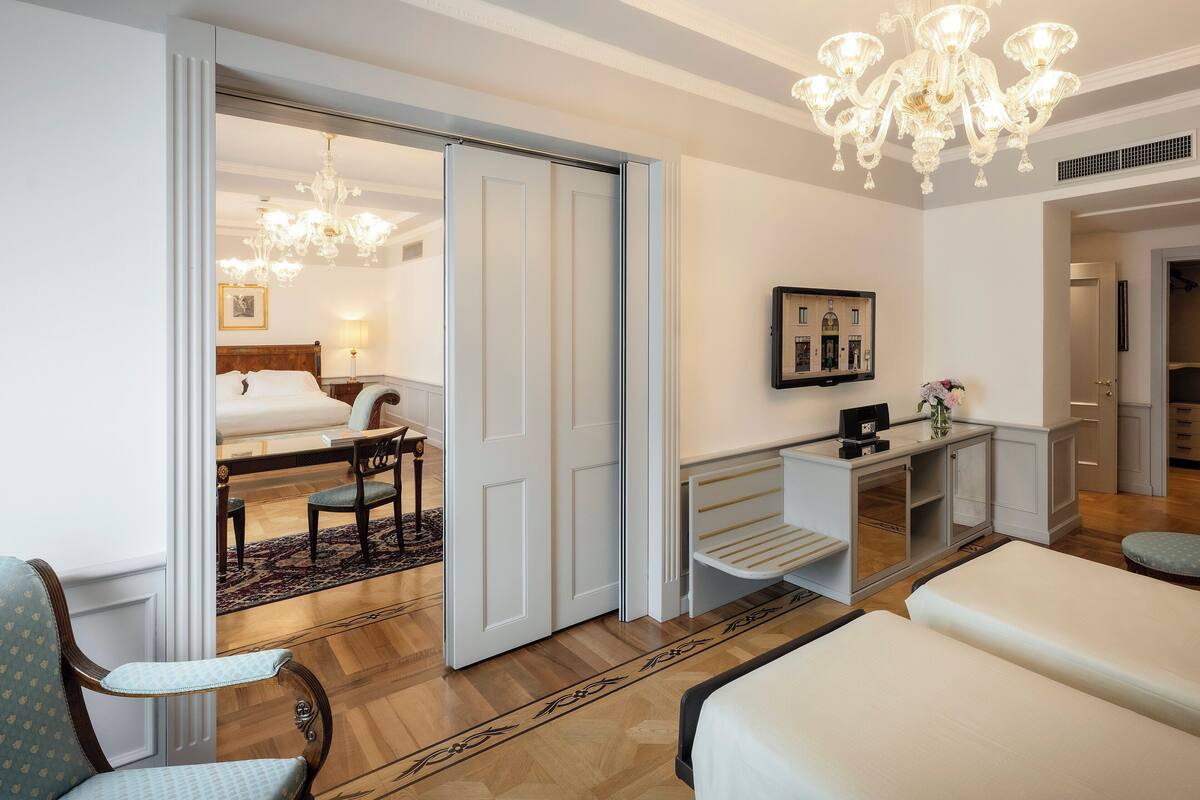 A hotel room with a bed, a desk, a chair, a chandelier, and a mirror on the wall.
