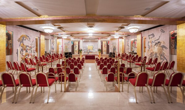 Un salón de conferencias con filas de sillas rojas y un escenario al frente.
