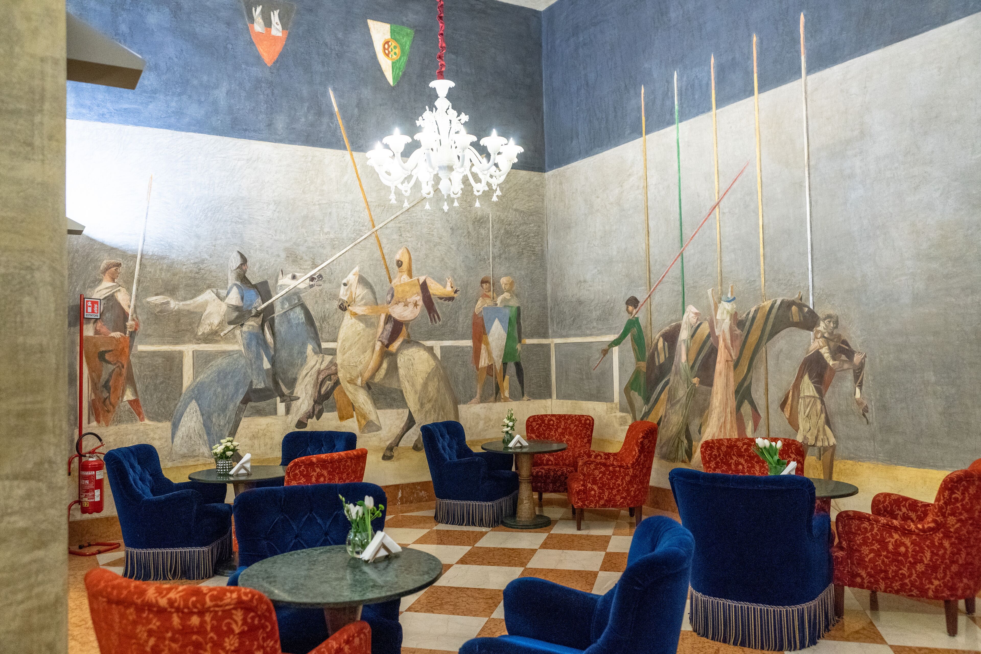 Un vestíbulo con piso a cuadros, sillones tapizados en azul y rojo, una araña y un mural que representa figuras históricas a caballo.