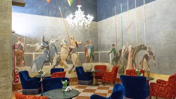 Un vestíbulo con piso a cuadros, sillones tapizados en azul y rojo, una araña y un mural que representa figuras históricas a caballo.