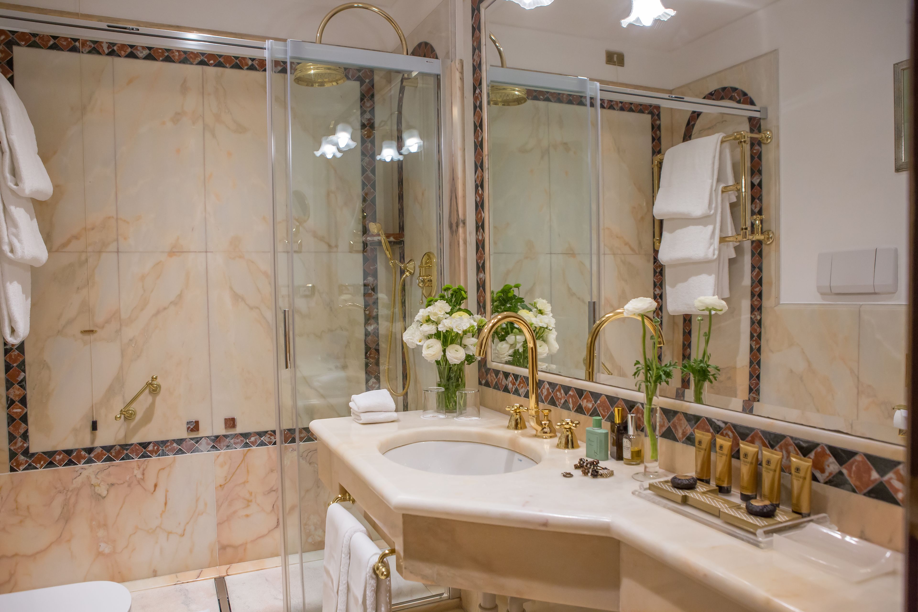 Ein Badezimmer mit einer gläsernen Duschkabine, einer mit Marmor belegten Wand, einem Waschbecken mit goldfarbenen Armaturen und einem Spiegel mit dekorativem Fliesenrahmen.