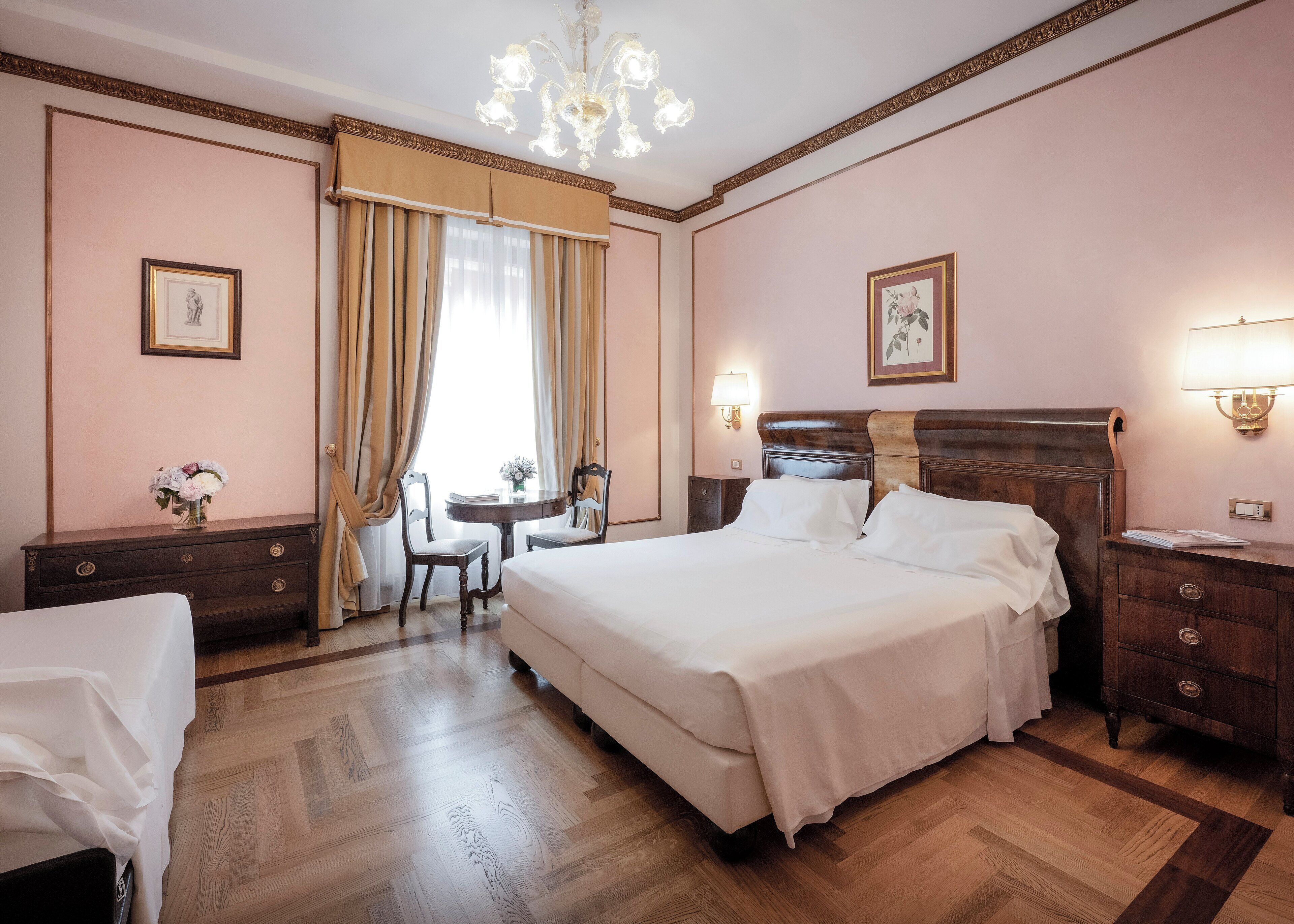 A hotel room with a large bed, a desk with a chair, a small table with a vase of flowers, and a nightstand with a lamp.