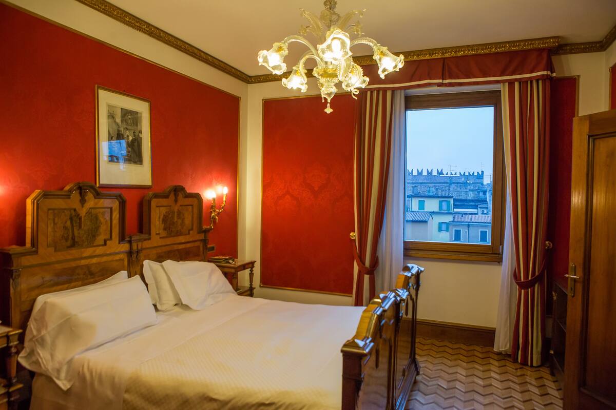 A hotel room with a large bed, a chandelier, a window with curtains, and a view of buildings outside.