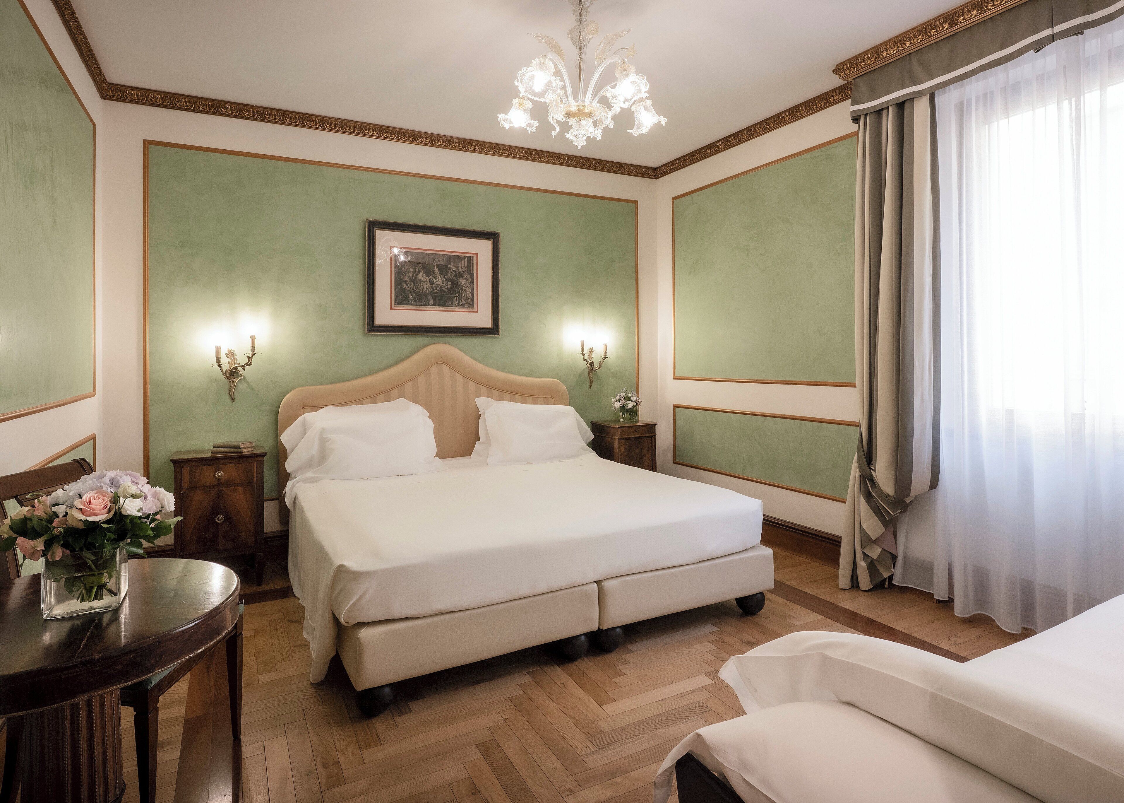 Un dormitorio ordenado con una cama grande, una mesita de noche con un florero, un cuadro en la pared y una araña.