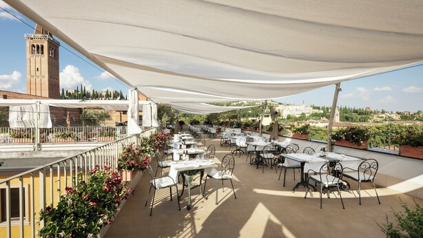 Una terraza al aire libre con mesas y sillas, un toldo blanco y vistas de un edificio histórico al fondo.