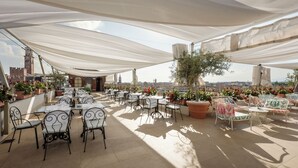 An outdoor terrace with tables and chairs, potted plants, and a view of the cityscape.