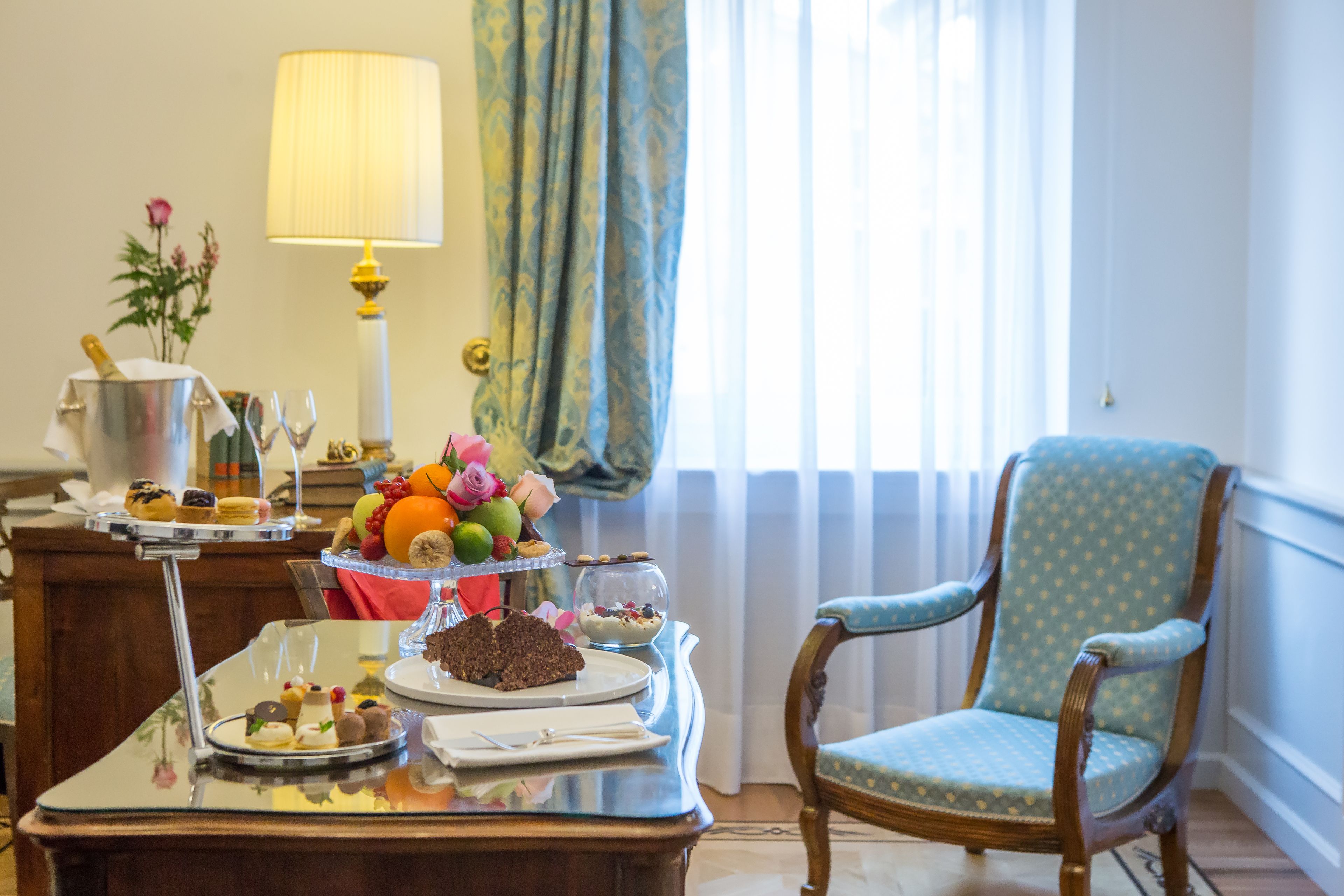 A dining table with a variety of desserts and fruits, a lamp, and a chair in a well-lit room with a window and curtains.