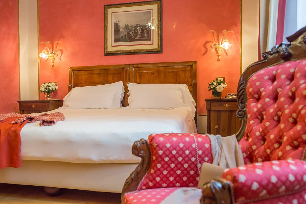 A bedroom with a large bed, two nightstands with lamps, a wooden headboard, a red upholstered chair, and a framed picture on the wall.