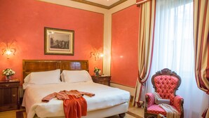 Una habitación de hotel con una cama grande, una mesita de noche de madera, un sillón rojo y un cuadro enmarcado en la pared.