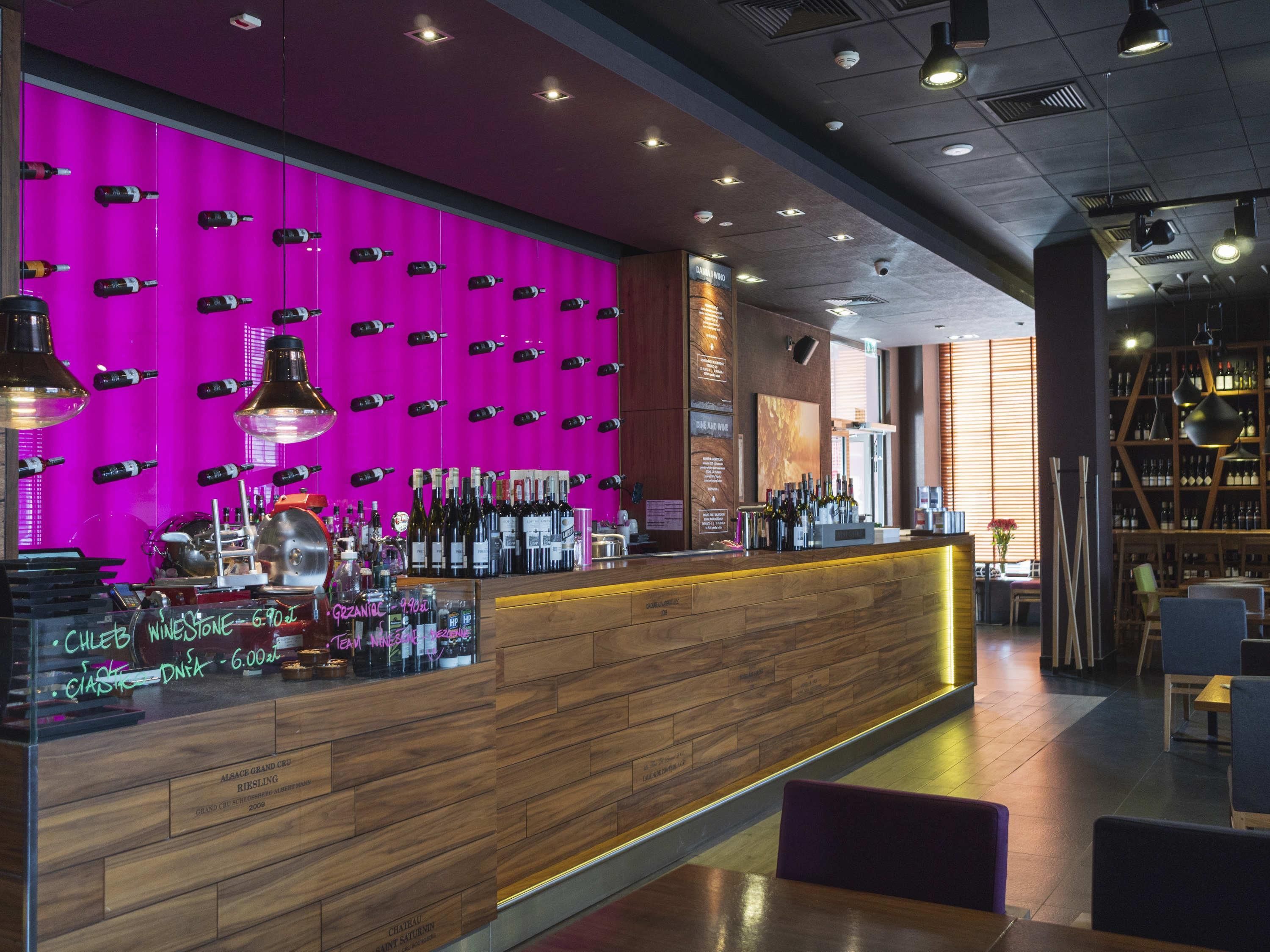 A bar area with a wooden counter, a pink wall with wine bottles, and a seating area with tables and chairs.