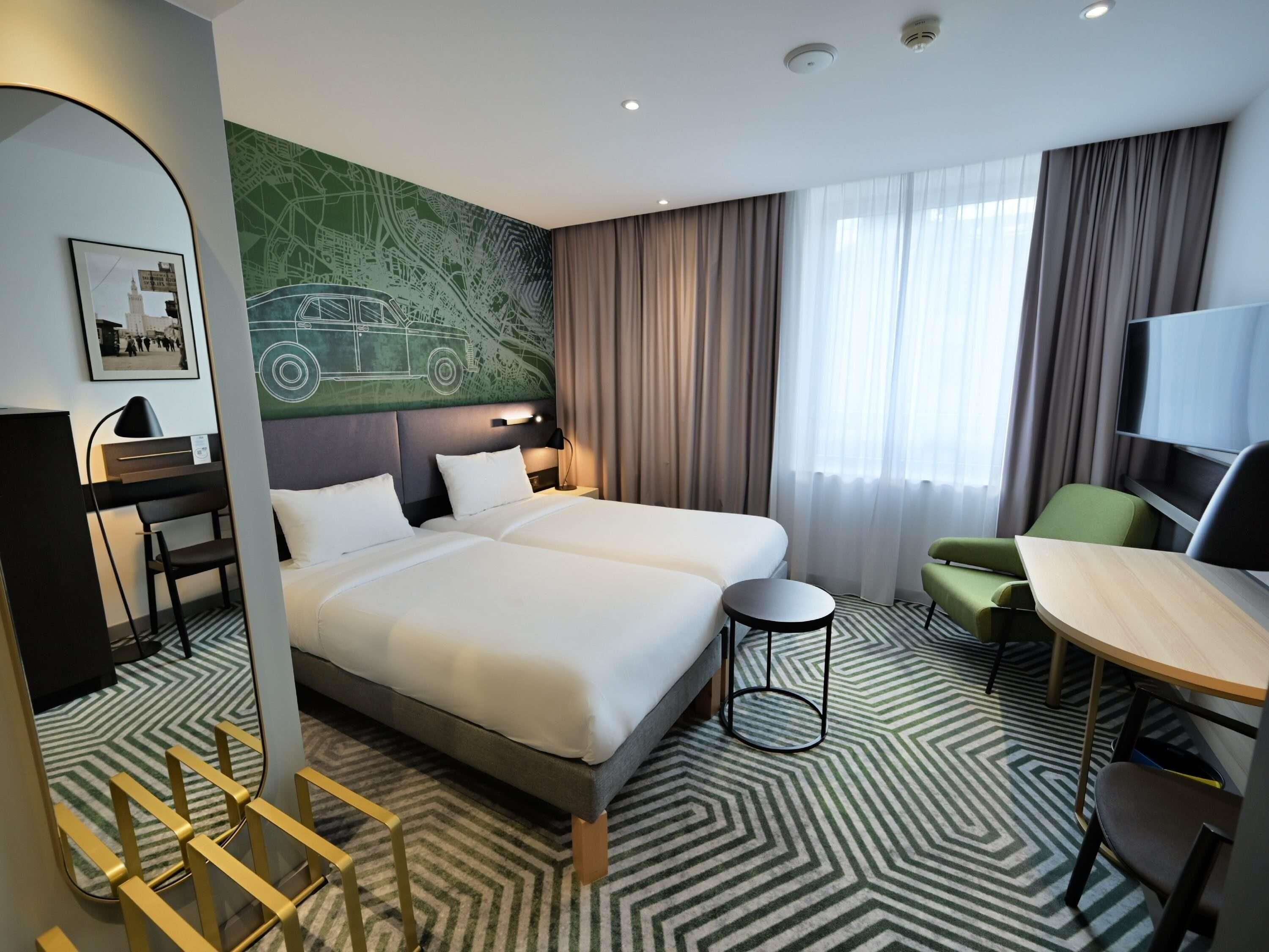 Habitació d'hotel amb un llit gran, una taula de treball, una cadira, un televisor i un mural a la paret.
