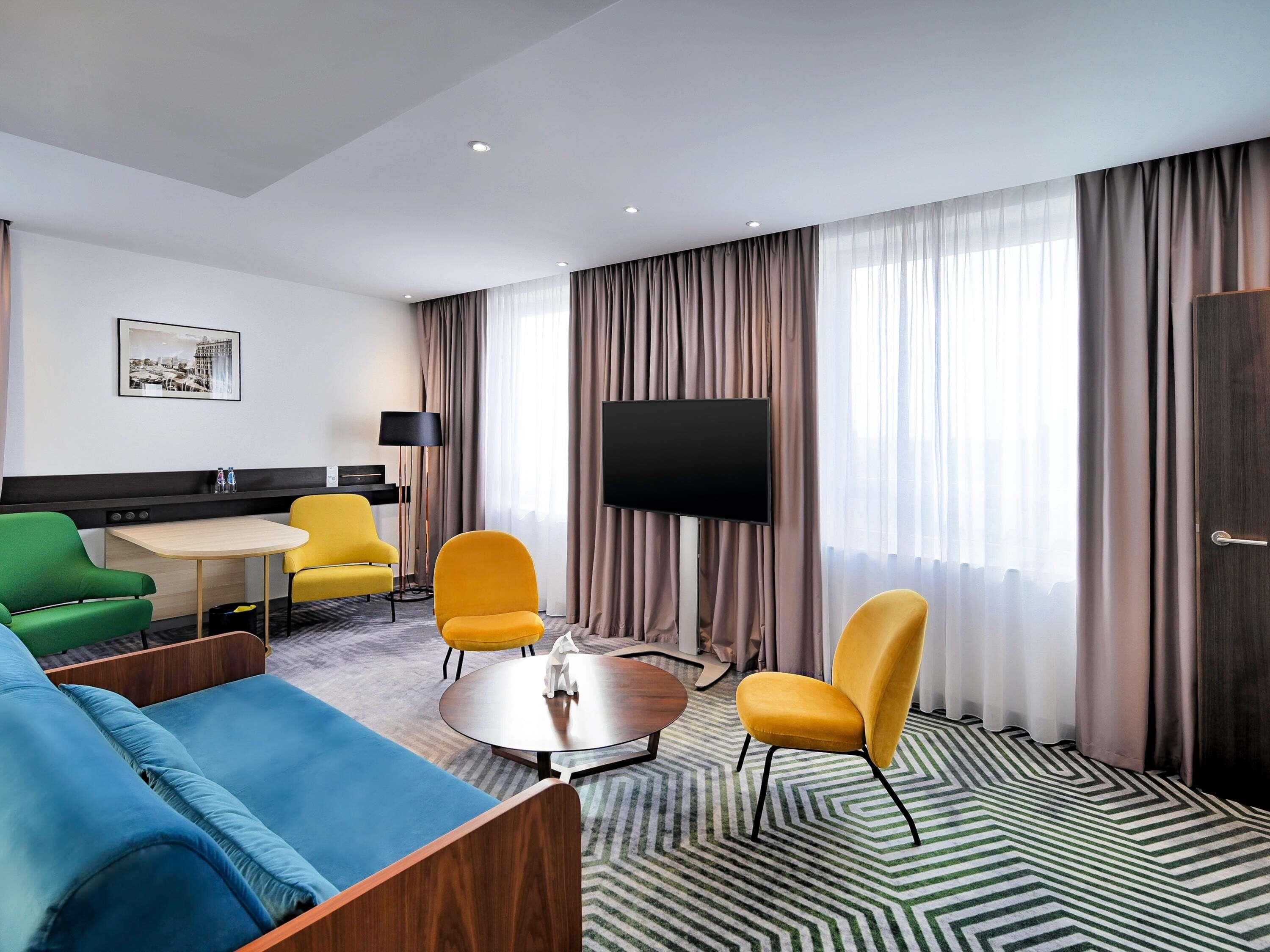 Habitación de hotel con sofá, sillas, una mesita, televisor y alfombra estampada.