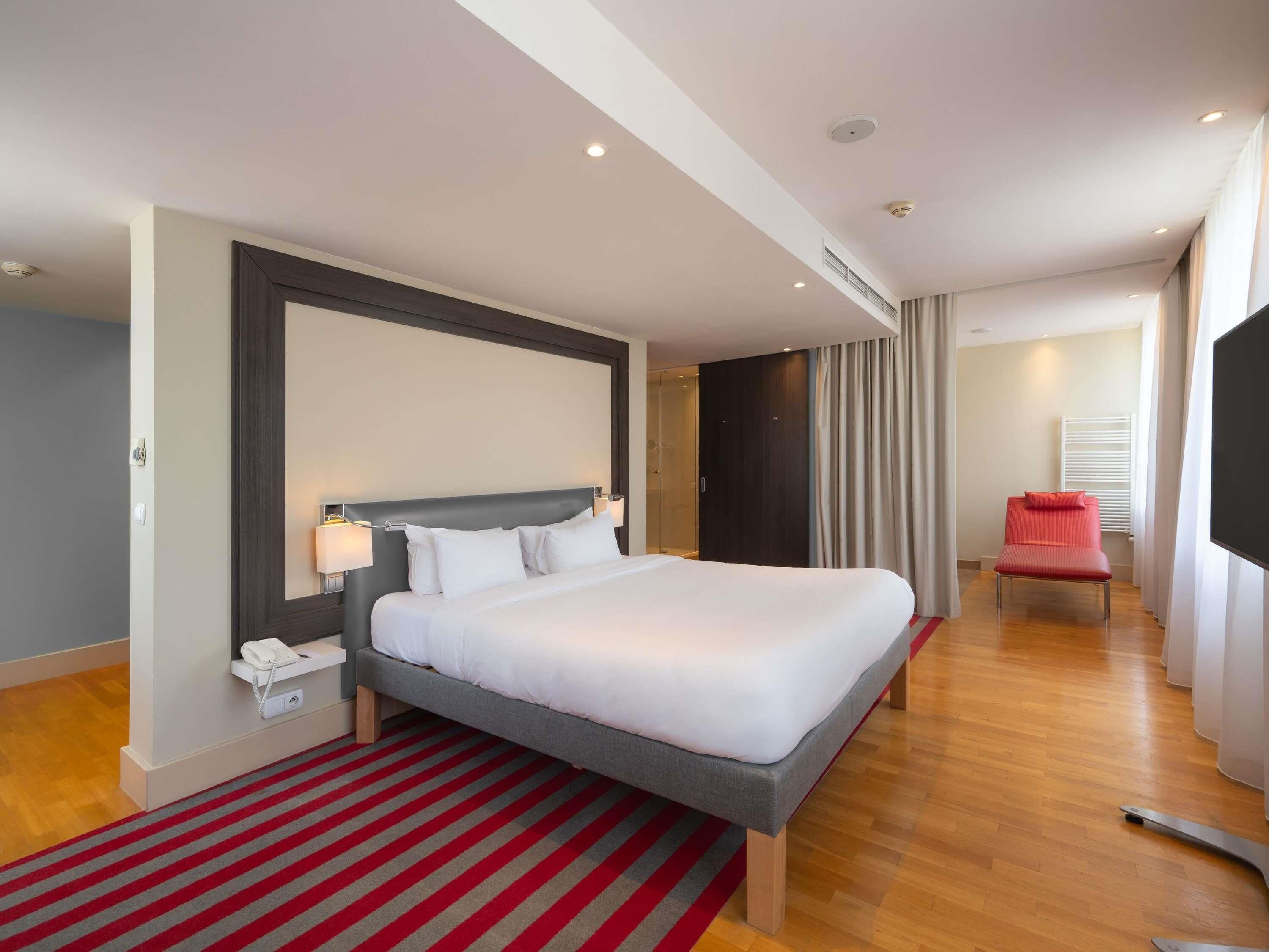 Une chambre d’hôtel moderne avec un grand lit, un tapis à rayures rouges, un fauteuil rouge et un téléviseur.