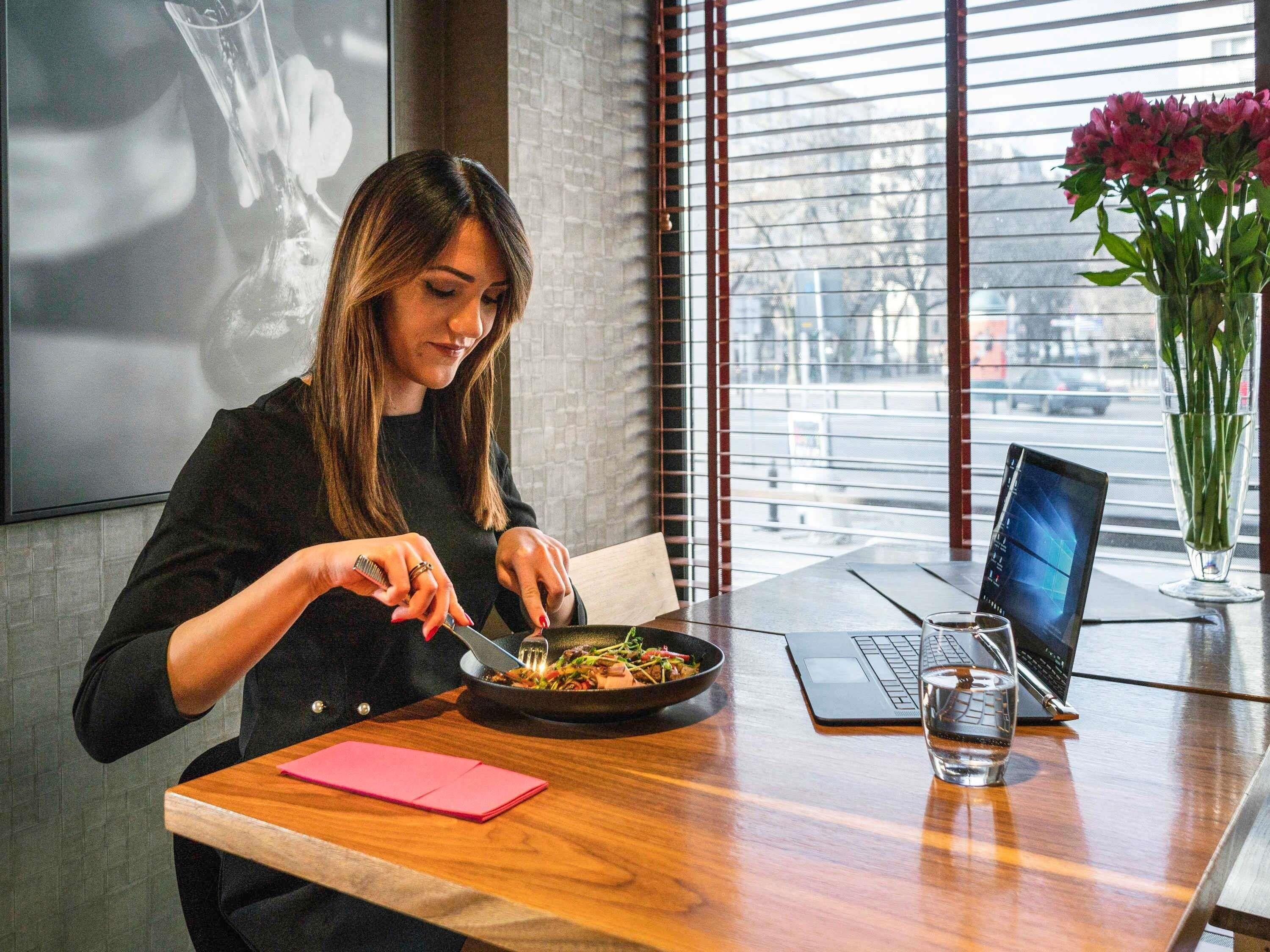 A person dining at a table with a laptop and a glass of water, with a window and a vase of flowers in the background.