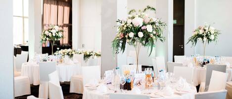 Un salón de banquetes con mesas redondas dispuestas para un evento formal, cada una con centros de mesa florales y cristalería.