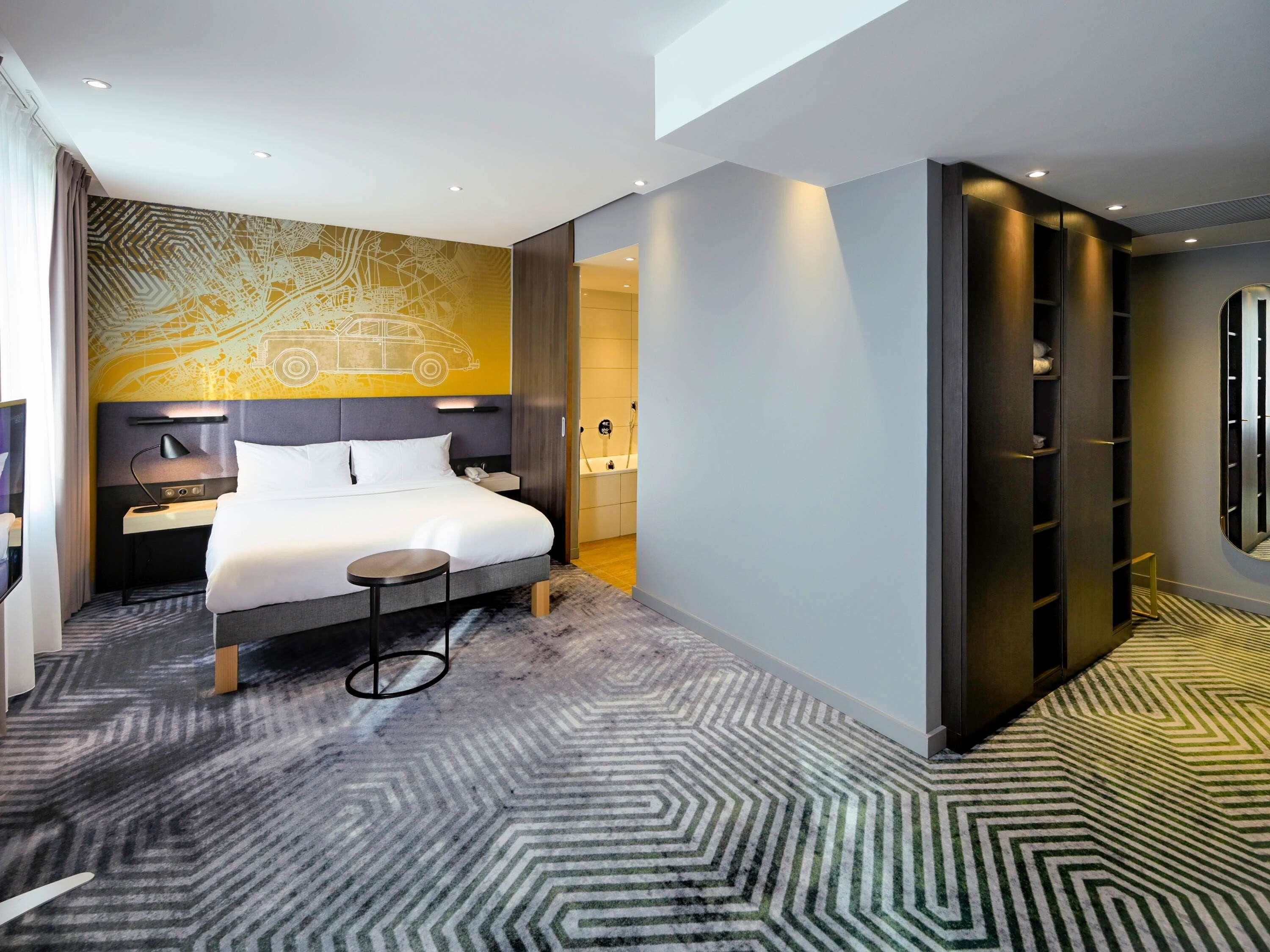 Une chambre d’hôtel moderne avec un grand lit, une petite table de chevet, un bureau et un dressing.