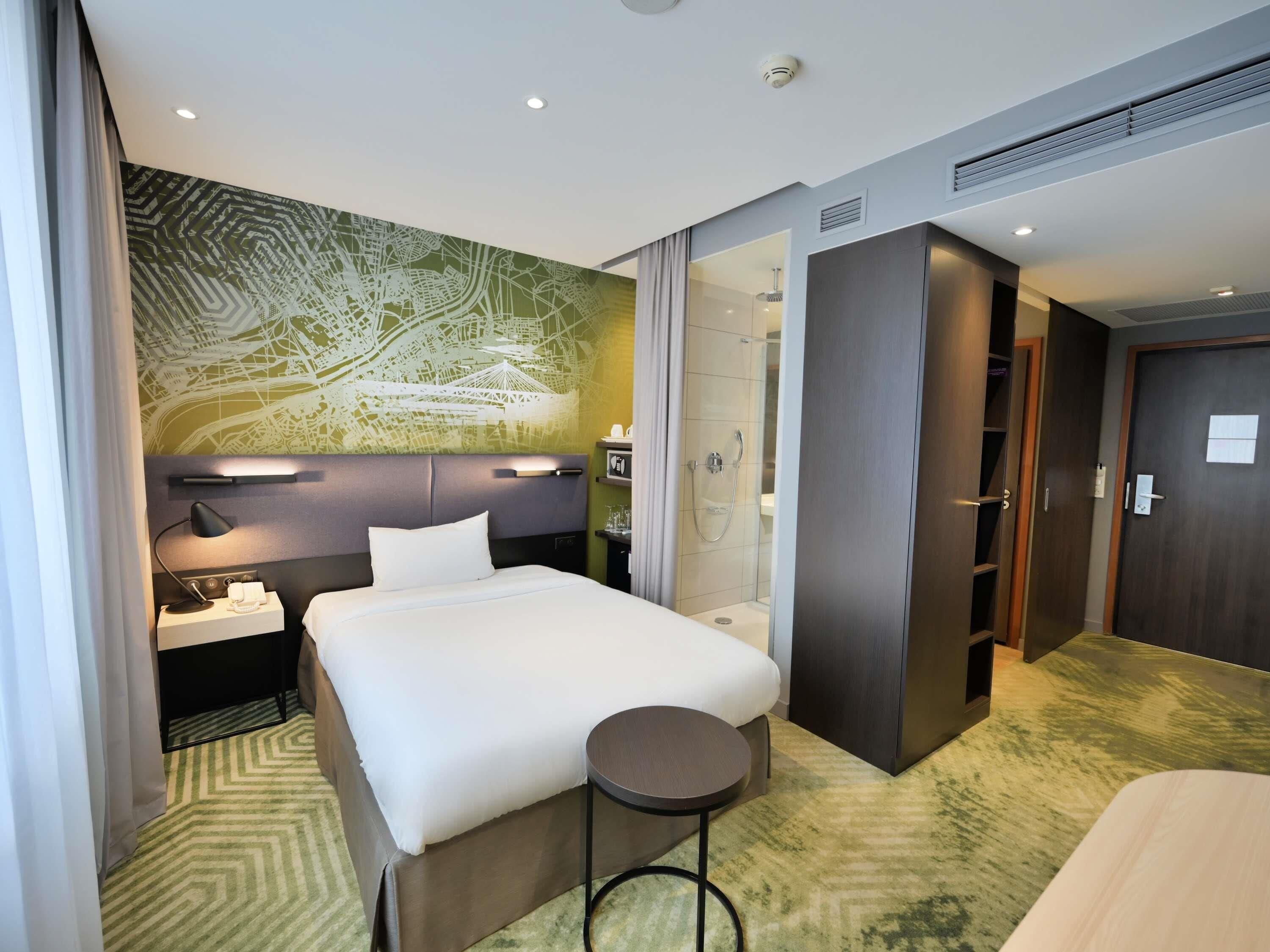 A modern hotel room with a bed, bedside table, lamp, and a shower area.