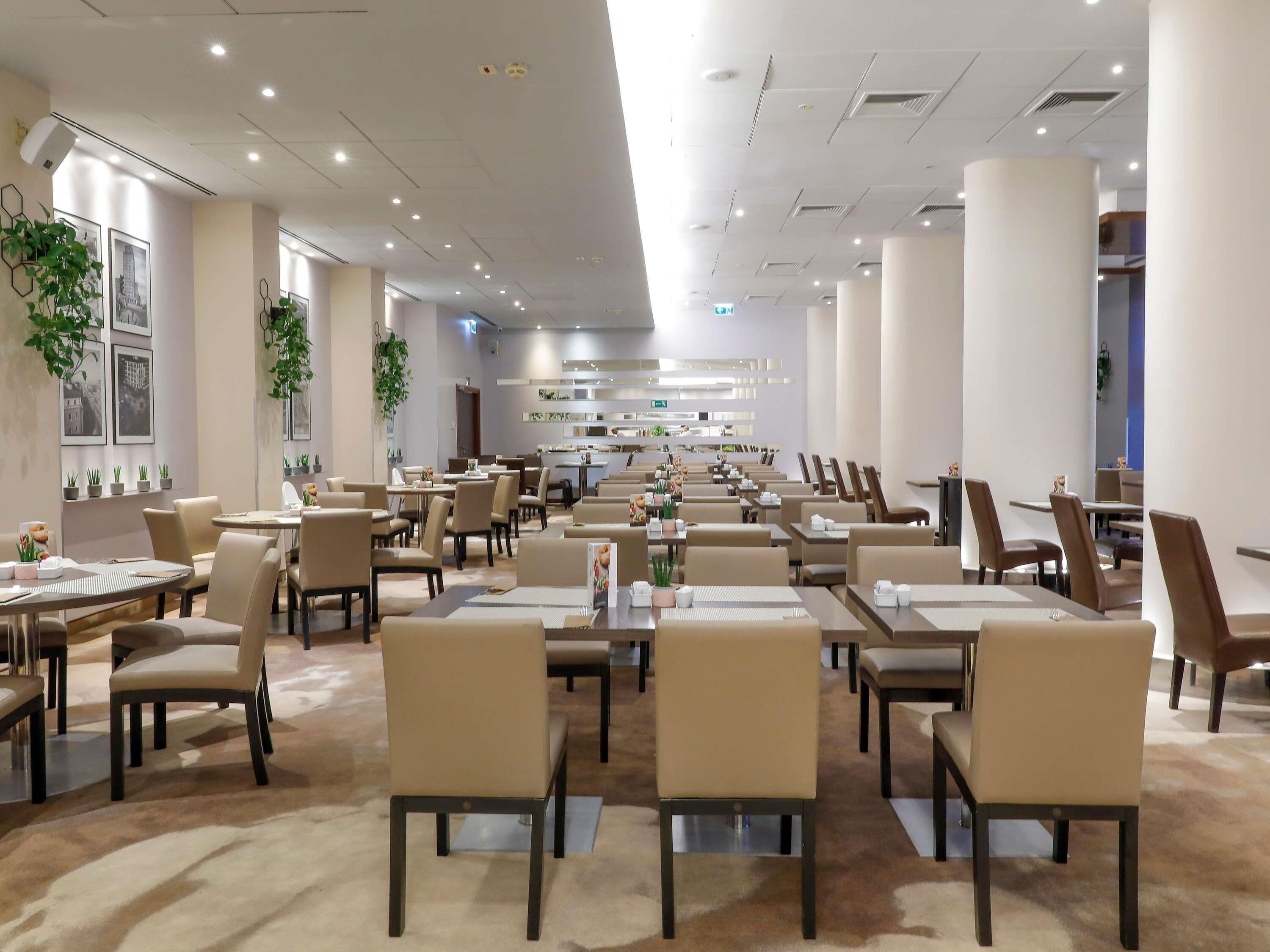 Un vaste espace de restauration d’hôtel, doté de nombreuses tables et chaises, d’un passage central et d’un design moderne au niveau du plafond.
