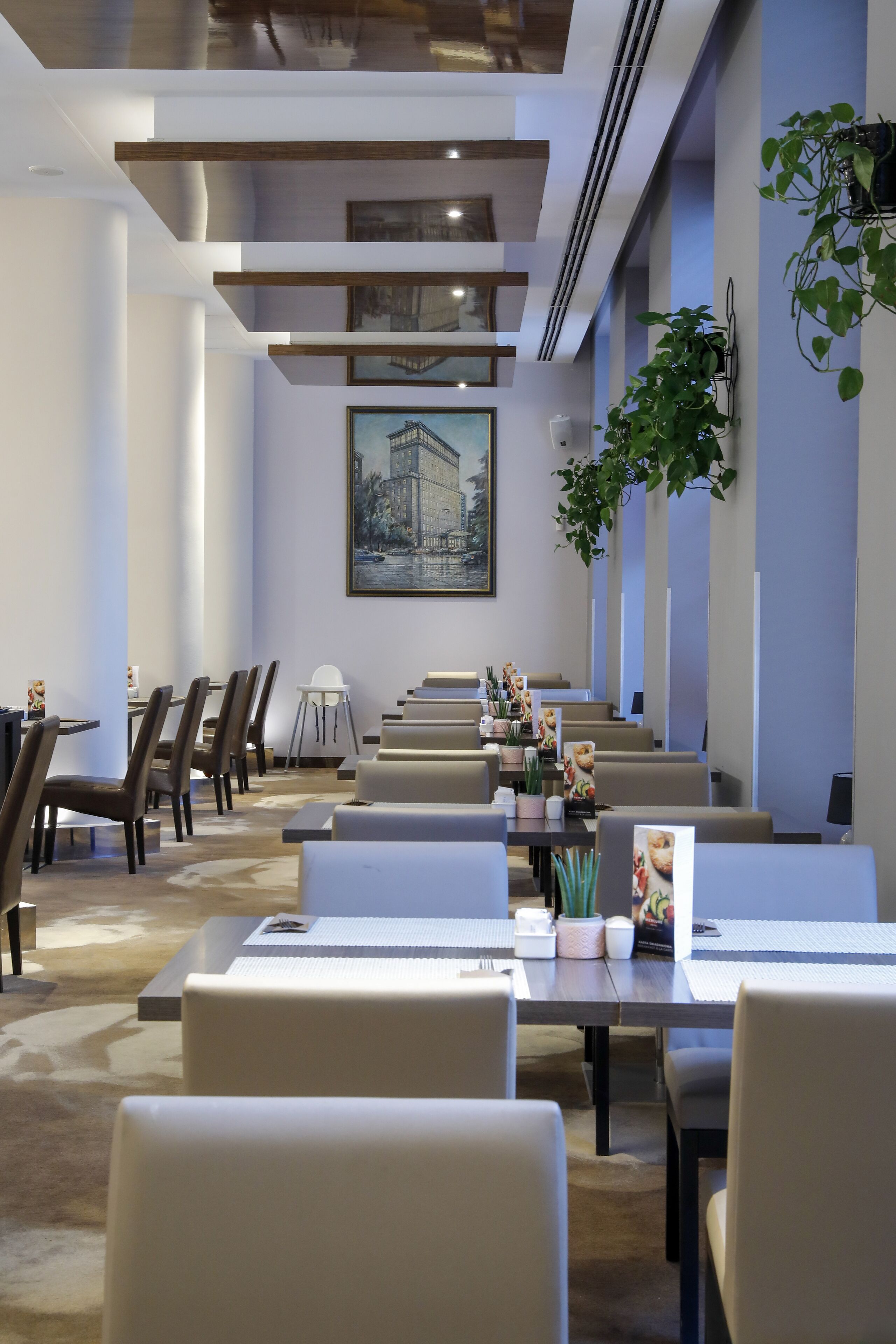 A modern hotel dining area with multiple tables and chairs, a large painting on the wall, and a potted plant.