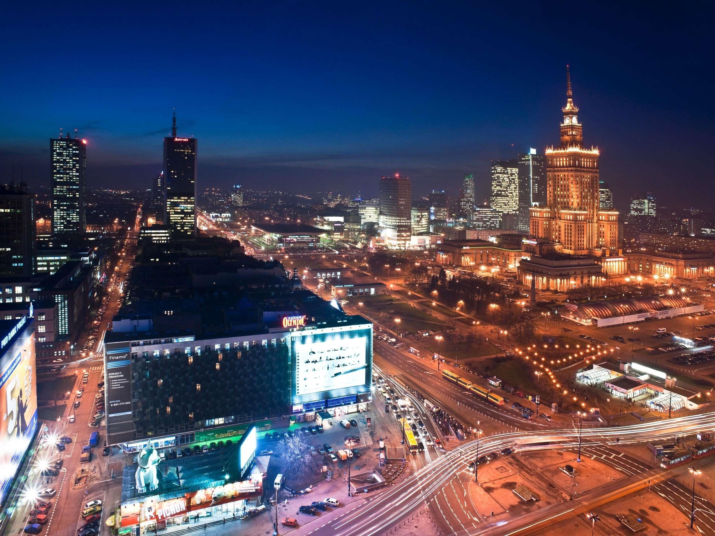 Une vue nocturne d’une ville, avec des bâtiments illuminés, une tour imposante et un grand panneau publicitaire.